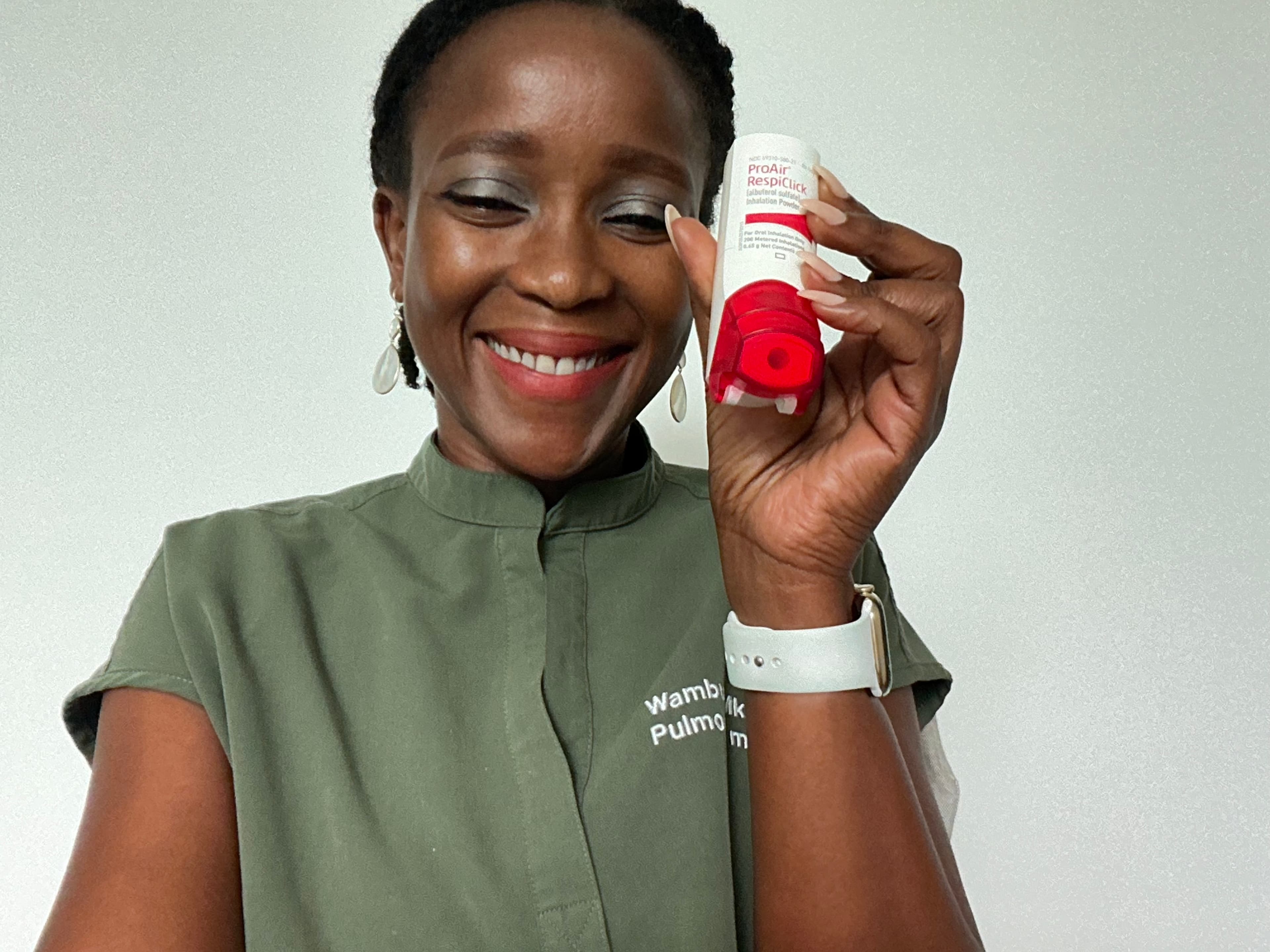 A smiling woman in green scrubs holds up a red and white ProAir RespiClick inhaler.